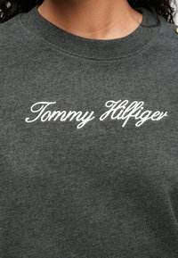 Dark gray sweatshirt with embroidered white "Tommy Hilfiger" logo, featuring a round neck, soft texture, and decorative shoulder buttons.