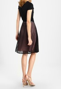 Svart topp i kombination med en genomskinlig kjol med rutmönster i mörkrosa, som har en figurnära midja och en utsvängd A-linjeform. Nakenfärgade högklackade skor.