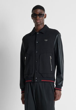 Uomo con giacca varsity nera con maniche in pelle, tasche con zip, polsini a righe rosse e occhiali da sole neri su uno sfondo grigio semplice.