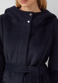 Marineblauer Fleece-Mantel mit Kapuze, Druckknöpfen und gebundenem Gürtel. Glatte Textur, strukturierte Nähte und Seitentaschen.