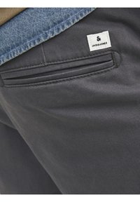 Graue Hose mit glatter Textur, ausgestattet mit einer seitlichen Tasche und einem kleinen weißen Etikett mit der Aufschrift "JACK & JONES". Oben ist ein Jeanshemd sichtbar.
