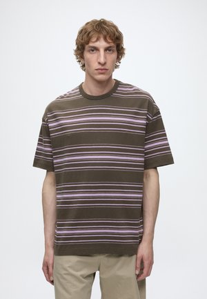 Jeune homme aux cheveux bruns clairs et bouclés, portant une chemise à manches courtes rayée olive et rose et un pantalon beige, debout sur fond blanc.