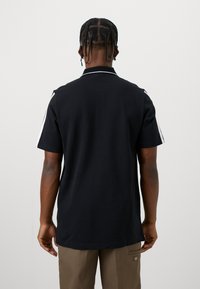 Muž s pletenými vlasy v černém polokošili s krátkým rukávem a bílými pruhy na rukávech a v hnědých kalhotách, otočený zády k bílému pozadí.