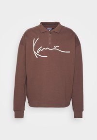 Sudadera marrón con cremallera y mangas largas, que presenta un logotipo blanco en el frente. Está hecha de una tela suave y texturizada.