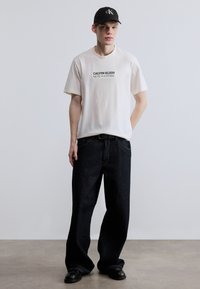 Λευκό βαμβακερό t-shirt με μαύρο γραφικό κείμενο, συνδυασμένο με σκούρο μπλε τζιν παντελόνι φαρδύ στο πόδι. Φορεμένο με μαύρο καπέλο και μαύρα παπούτσια.