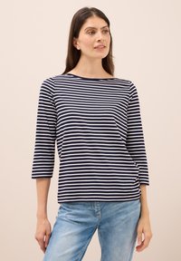 Marineblau und weiß gestreiftes T-Shirt mit drei Viertelärmeln und Bootsausschnitt. Aus weichem Stoff und mit lockerem Sitz. Kombiniert mit hellblauen Jeans.