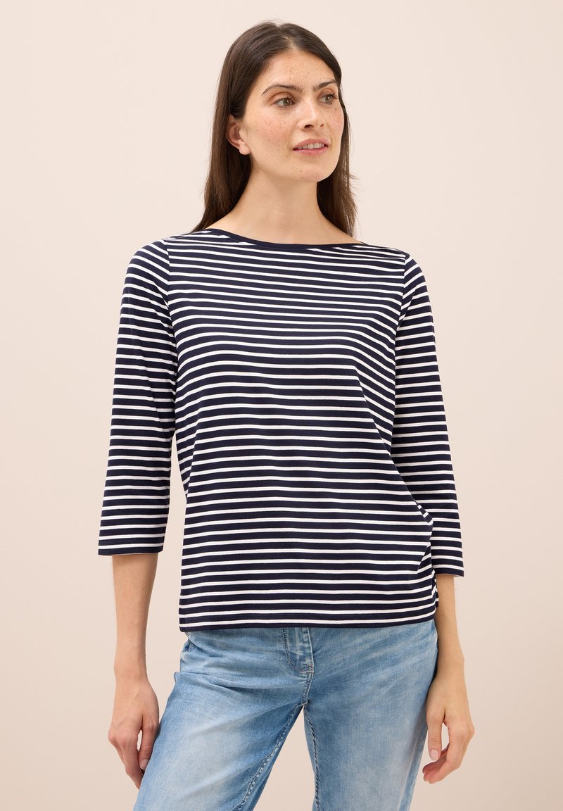 Marineblau und weiß gestreiftes T-Shirt mit drei Viertelärmeln und Bootsausschnitt. Aus weichem Stoff und mit lockerem Sitz. Kombiniert mit hellblauen Jeans.