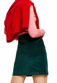 Groene corduroy rok met steekzakken aan de voorkant en een nauwsluitende tailleband, gecombineerd met een rood gebreide trui over een roze shirt met lange mouwen.