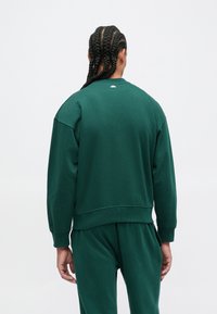 Pessoa com cabelo trançado de costas, vestindo um sweatshirt verde escuro e calças combinando, contra um fundo claro e simples.