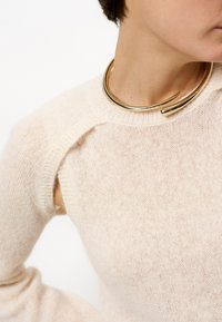 Cremfarbener Strickpullover mit lockerer Passform, ausgestattet mit einem gerippten Halsausschnitt und einem einzigartigen Ausschnitt an der Schulter. Goldene Metall-Choker-Akzent.