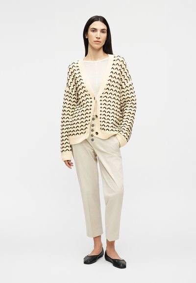 Γυναίκα που φοράει κρεμ ζακέτα με σχέδια, μπεζ παντελόνι ψηλόμεσο με κουμπιά, λευκό τοπ και μαύρες παντόφλες, στέκεται με το ένα χέρι στην τσέπη.