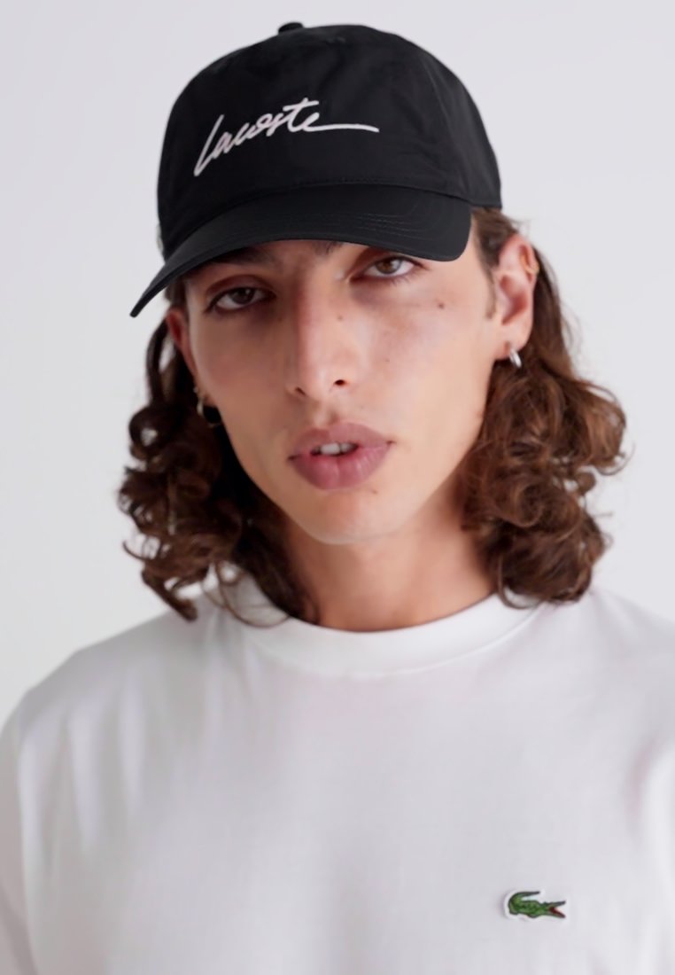 Casquette noire avec du texte manuscrit blanc ; portée avec un t-shirt blanc uni arborant un petit logo de crocodile vert sur la poitrine.