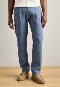Lichtblauwe linnen broek met een ontspannen pasvorm en voorplooien, gecombineerd met beige moccasin-stijl schoenen en een gestructureerd crème top.