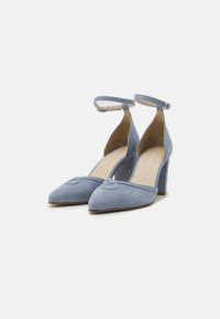 Zapatos de tacón bajo de ante azul claro, con punta afilada, tiras en el tobillo y un detalle decorativo en forma de corazón en la puntera. Textura suave con un ligero brillo.