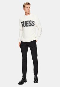 Maglione bianco con logo "GUESS" in paillettes nere sul petto, abbinato a pantaloni neri e scarpe nere. Il modello è in piedi con un'espressione neutra.
