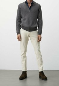 Grauer Reißverschluss-Pullover mit strukturiertem Design, cremefarbene Hose und braune Schnürstiefel. Das Outfit zeigt eine figurbetonte Silhouette.
