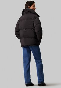 Zwarte pufferjack met hoge kraag, gestikt ontwerp en volumineuze silhouet, gecombineerd met blauwe spijkerbroek en zwarte schoenen.