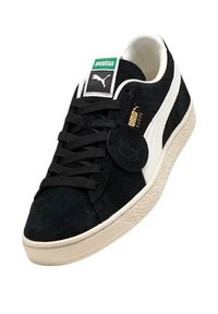 Sneaker Puma Suede nera con striscia bianca, suola beige, lacci neri e logo verde sulla linguetta, angolata per mostrare il lato e la parte superiore.