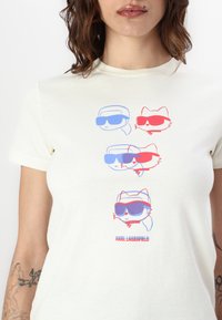 Krémové bavlnené tričko s štyrmi kreslenými postavičkami nosiacimi slnečné okuliare v červenej a modrej farbe, s nápisom "KARL LAGERFELD" na spodnej strane.