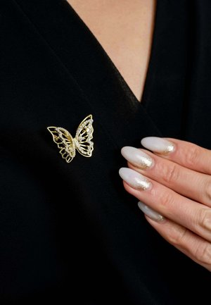 Gold butterfly brooch with intricate cutouts and crystal accents, placed on a black garment. Long, light nails with a metallic finish visible.