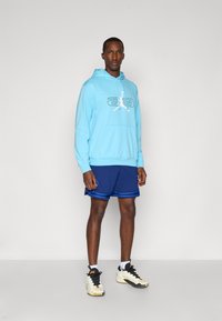 Sudadera azul claro con un logo blanco, combinada con pantalones cortos azul marino con rayas azules. Zapatillas deportivas en blanco y negro. Fondo minimalista.