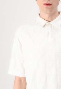 Hvit polo skjorte med et teksturert mønster med hevede bokstaver og former, korte ermer og en klassisk krage med knapper.