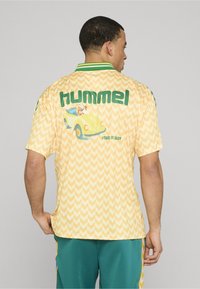 Lühikeste varrukatega kollane särk tikandmustriga, rohelise kraega ning tagaosas Hummeli logo koos auto multikajoonise ja fraasiga "#TAKE IT EASY".