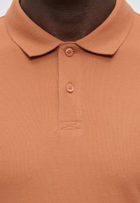 Άτομο με σκοτεινό δέρμα που φοράει ανοιχτό καφέ πόλο μπλούζα με γιακά και δύο καφέ κουμπιά κουμπωμένα στη λαιμόκοψη.