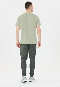 Man som bär en ljusgrön kortärmad skjorta, mörkgrå byxor med förstärkta underben och vita sneakers, vänd bort från kameran mot en vit bakgrund.