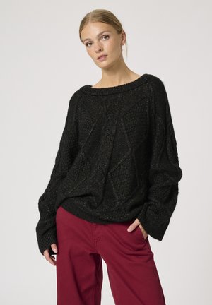 Sort strikket sweater med løs pasform, strukturerede diamantmønstre, bred halsudskæring og ribbede manchetter, parret med bordeauxfarvede bukser.