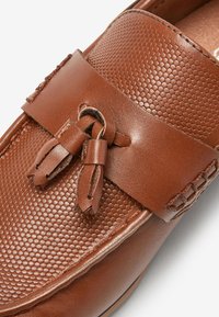 Chaussure slip-on en cuir marron avec un design texturé en nid d'abeille et des pampilles en cuir. Accents lisses sur le dessus avec des coutures détaillées sur les côtés.