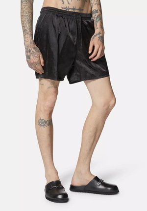 Costumi da bagno neri con un motivo sottile, vita elastica e tasche laterali, abbinati a sandali neri con una fascia decorativa.
