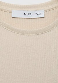 Beige geripptes Stricktop mit Rundhalsausschnitt und strukturiertem Stoff. Das Etikett trägt die Aufschrift "MNG" mit Größenangaben. Einfaches Design, keine weiteren Details.