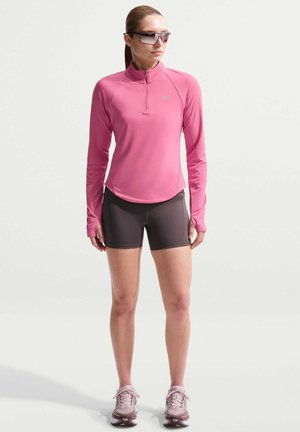 Femme portant un haut de sport rose à manches longues, un short gris foncé, des chaussures de course roses et des lunettes de soleil, debout devant un fond clair uni.