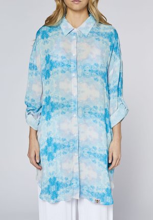 Woman wearing long, buttoned, collared shirt with blue and white abstract cloud pattern and rolled-up sleeves, paired with white pants.