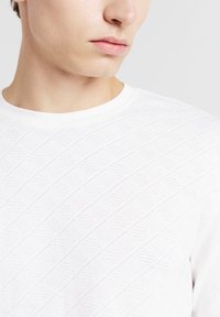 Primo piano di una persona che indossa una maglietta bianca a collo rotondo con texture, focalizzandosi sulla parte inferiore del volto e sulla parte superiore del torso.