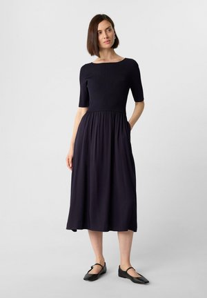 Navyblaues Kleid mit einem gerippten Oberteil und kurzen Ärmeln, das in einen fließenden Rock übergeht. Verfügt über seitliche Taschen und einen runden Ausschnitt.