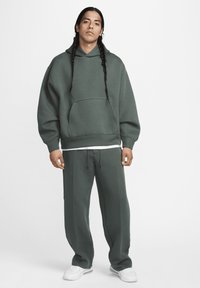 Sweatshirt verde-escuro com bolso canguru e capuz com cordão, combinado com calças de moletão justas a condizer e ténis brancos.
