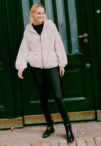 Hoodie zippé en tissu doux de couleur rose clair avec une capuche, de grandes poches et des poignets resserrés, associé à un jean slim noir et des bottes chelsea noires.