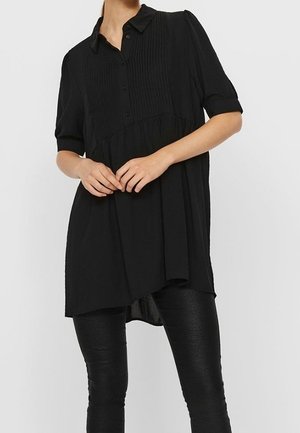 Kobieta ubrana w luźną czarną koszulę na krótki rękaw z zapięciem na guziki oraz czarne, teksturowane, obcisłe spodnie, stojąca ze splecionymi rękami przed sobą.