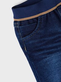 Jeans de mezclilla azul oscuro con una amplia cintura elástica en tono navy, decorada con una costura dorada. Incluye detalles en los bolsillos frontales.