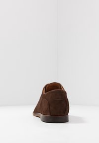 Scarpa in suede marrone con texture liscia, tacco basso, punta arrotondata e dettagli di cucitura minimi, su uno sfondo bianco semplice.