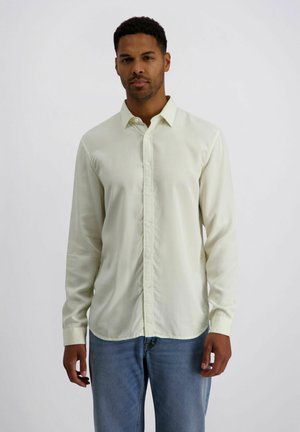 Mann trägt ein hellbeige Hemd mit langen Ärmeln und Knöpfen sowie blaue Jeans, steht vor einem schlichten weißen Hintergrund.