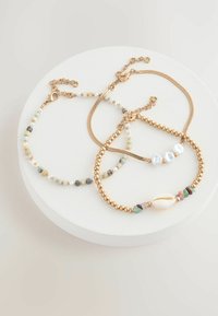Tre braccialetti su un supporto rotondo bianco: due presentano catene d'oro con perline e uno include un accento di conchiglia insieme a pietre colorate e perle.