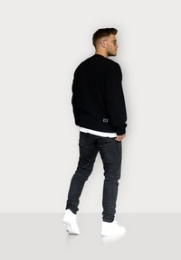 Schwarzer Strickpullover mit geripptem Saum, über einem weißen Hemd getragen, kombiniert mit dunklen Jeans und weißen Sneakers. Mit einem Logo am Saum.