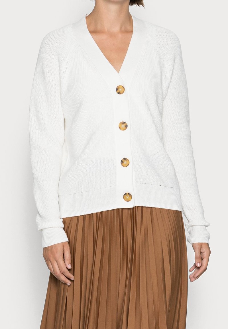 Cardigan bianco a maglia con scollo a V, caratterizzato da grandi bottoni rotondi dorati e polsini a coste. Abbinato a una gonna marrone plissettata.