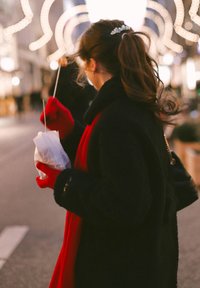 Zwarte fleece jas, rode sjaal en handschoenen. Houdt een ingepakt item vast met een rietje. Zachte textuur en warme kleuren, met decoratieve lichten op de achtergrond.