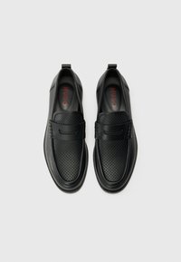 Svarte slip-on sko med perforert lær, en jevn tådesign og en kontrasterende rød logo på innersålen. Rund gummisåle.