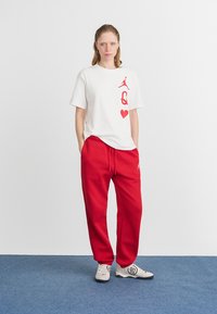 T-shirt branca de algodão com gráfico em vermelho e design de coração, combinada com calças de treino vermelhas largas. Os sapatos são cinzentos com um logo preto.