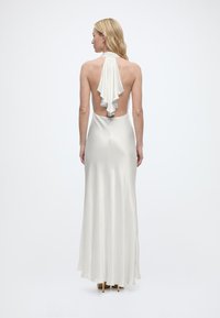 Γυναίκα με μακριά ξανθά μαλλιά που φοράει λευκό φόρεμα χωρίς πλάτη με λεπτομέρεια από ρέον ύφασμα, στέκεται μπροστά σε ανοιχτό φόντο.